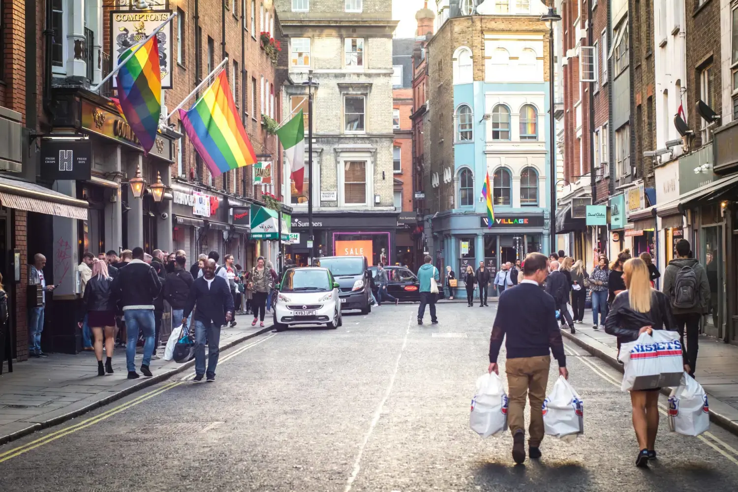 Old Compton Street in Soho on a busy afternoon, with rainbow flags above the pubs and shoppers in the road.