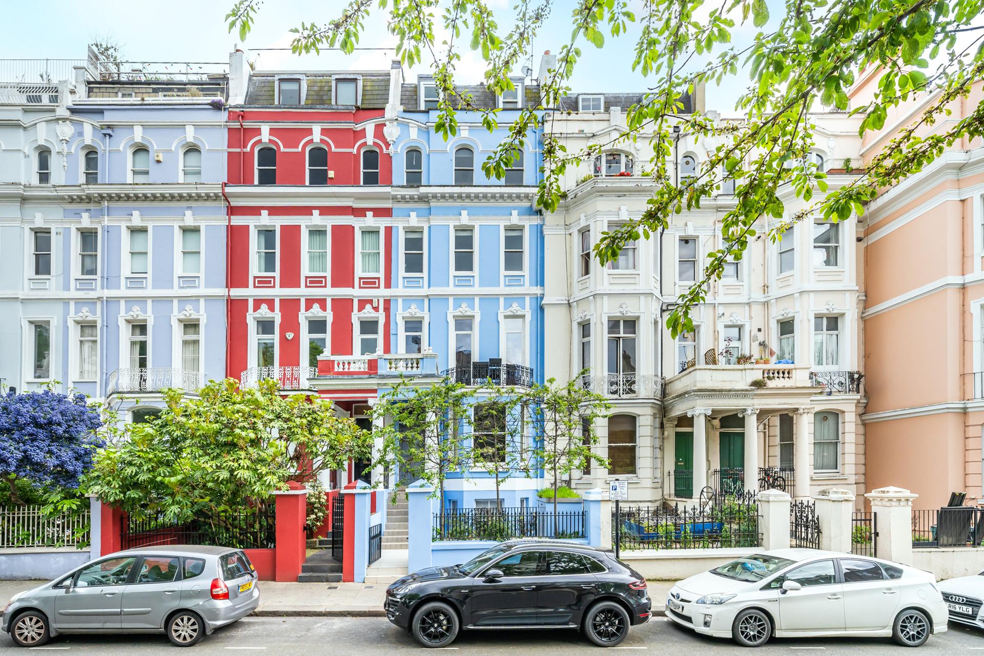 A Notting Hill terrace of painted stucco houses with raised ground floors, railings and cars parked nose to tail along the kerb.