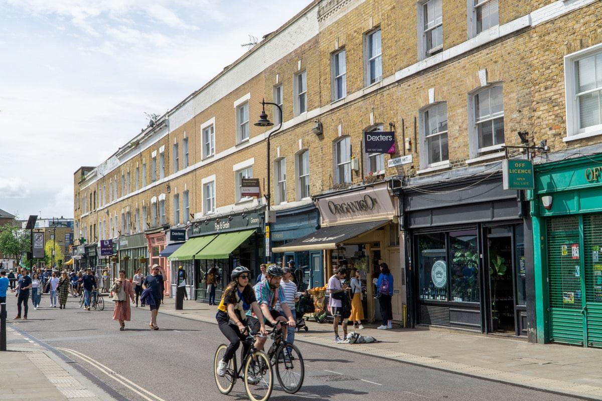 Broadway Market in Hackney on a busy afternoon, yellow brick terraces with shopfronts below and cyclists in the road.